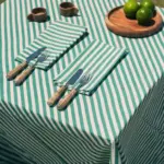 Mantel de rayas verdes y blancas de Cassai con servilletas a juego, cubiertos de fibras naturales y limas sobre mesa de inspiración mediterránea.
