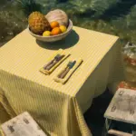 Mantel de rayas amarillas y blancas de Cassai sobre mesa junto al mar con frutero tropical y cubiertos, ideal para decoración mediterránea de verano.
