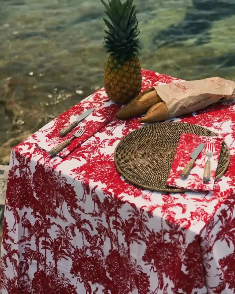 Detalle del mantel Garza Roja de Cassai con estampado de garzas y palmeras, pan artesanal y vajilla sobre una mesa mediterránea.