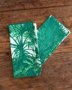 Servilletas de tela con estampado de palmeras verdes sobre fondo blanco, dobladas sobre mesa de madera rústica