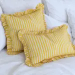 Fundas de cojín estampadas a rayas amarillas y blancas con acabado de volantes, combinadas con textiles blancos en una decoración luminosa y mediterránea.