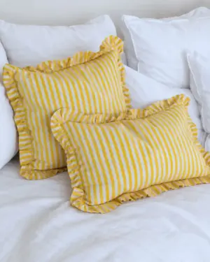 Fundas de cojín estampadas a rayas amarillas y blancas con acabado de volantes, combinadas con textiles blancos en una decoración luminosa y mediterránea.