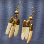 Par de pendientes colgantes artesanales compuestos por tres piezas alargadas de nácar color marfil con las puntas chapadas en oro, suspendidas de una bola dorada texturizada y un gancho tipo francés en tono dorado, sobre fondo azul.