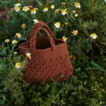 Bolso crochet color chocolate artesanal colocado entre flores silvestres con asas tejidas y textura detallada
