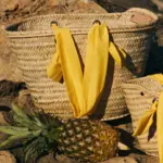 Cesta de palmito grande con asas amarillas sobre rocas junto a una piña, bolso artesanal estilo mediterráneo.