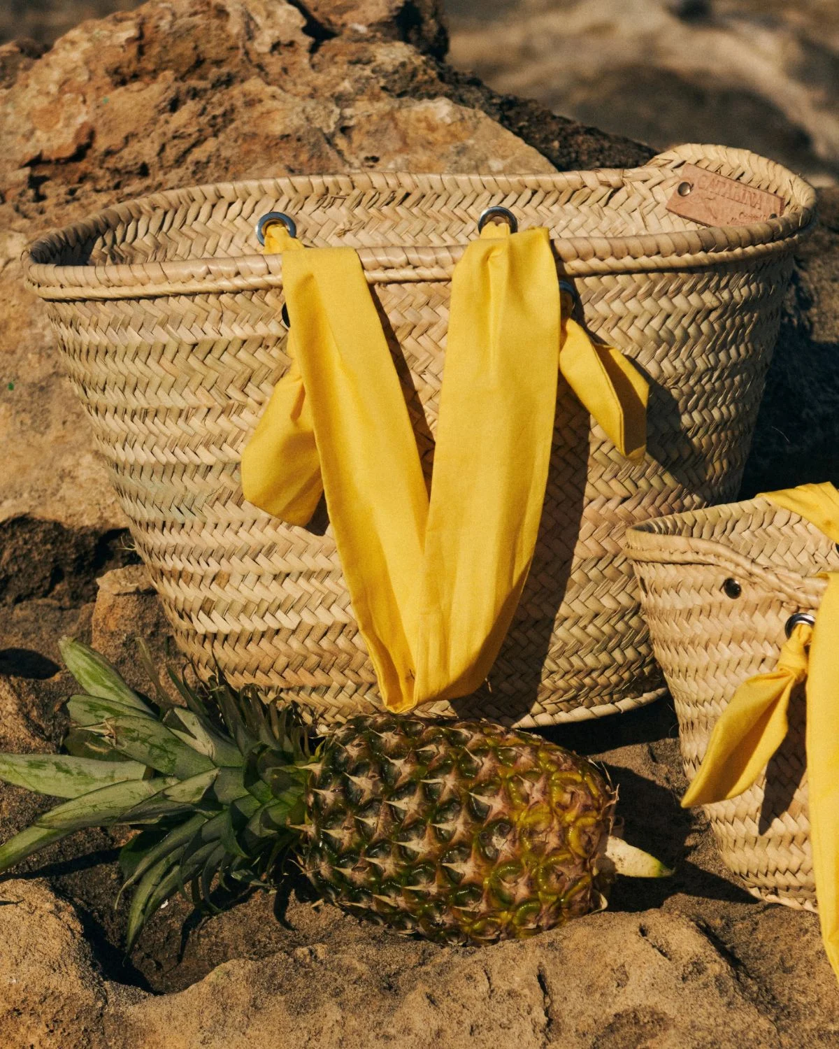Cesta de palmito grande con asas amarillas sobre rocas junto a una piña, bolso artesanal estilo mediterráneo.