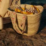 Cesta grande de palmito con asas de estampado floral sobre roca junto al mar en estilo mediterráneo