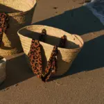 Cesta de palmito mediana con acabado estilo rafia y asas animal print sobre arena de playa, bolso de verano natural hecho a mano.