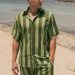Modelo vistiendo la Camisa Unisex Rayas Mediterráneas Verde de Cassai Home en la playa, una camisa oversize de verano con rayas verdes e inspiración mediterránea.