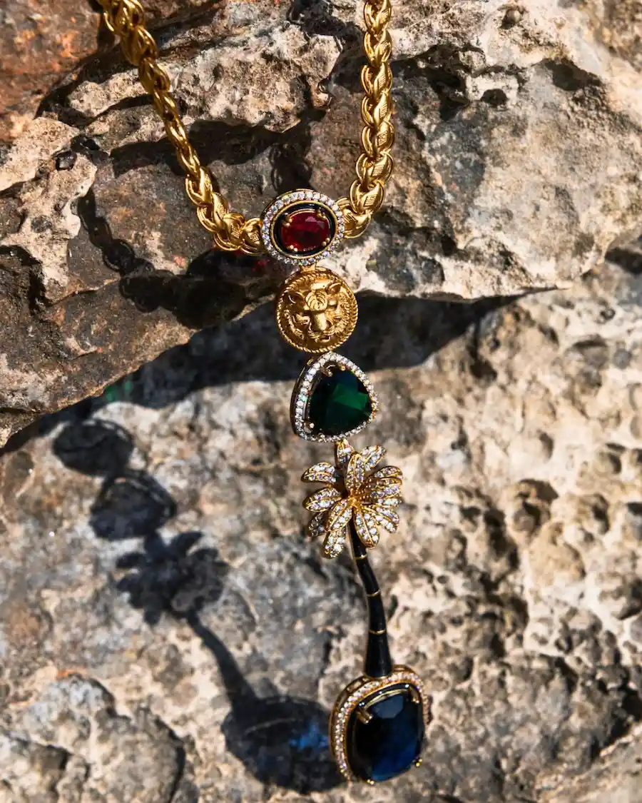 El Collar Oasis Imperial Azul Mediterráneo Cassai Home combina una cadena de eslabones dorados con cristales de colores, un medallón con cabeza de león y una palmera ornamentada, inspirándose en la riqueza y el lujo del Mediterráneo.