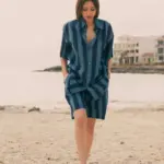 Modelo caminando por la playa con la Bermuda Unisex Rayas Mediterráneas azul marino de Cassai Home, una bermuda de verano de corte relajado e inspiración mediterránea.