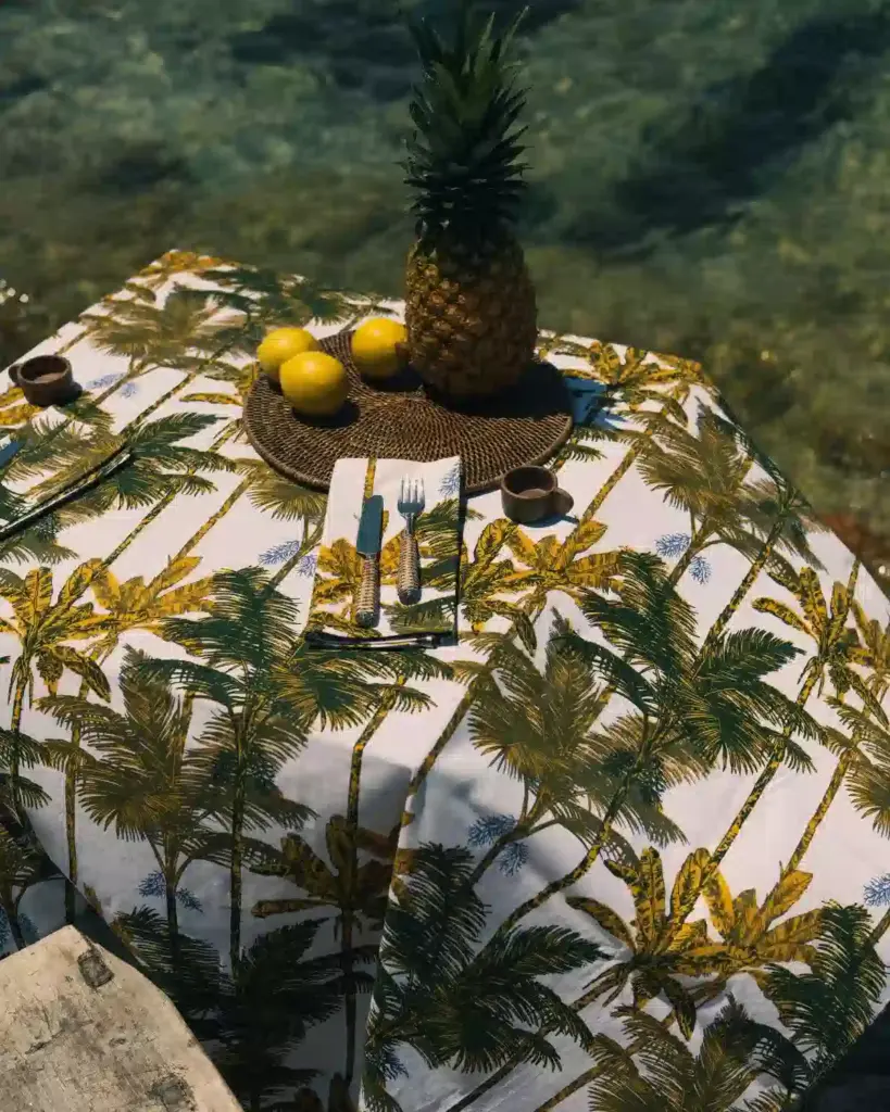 Mantel de algodón con estampado de palmeras mostaza de Cassai sobre una mesa decorada junto al mar Mediterráneo.