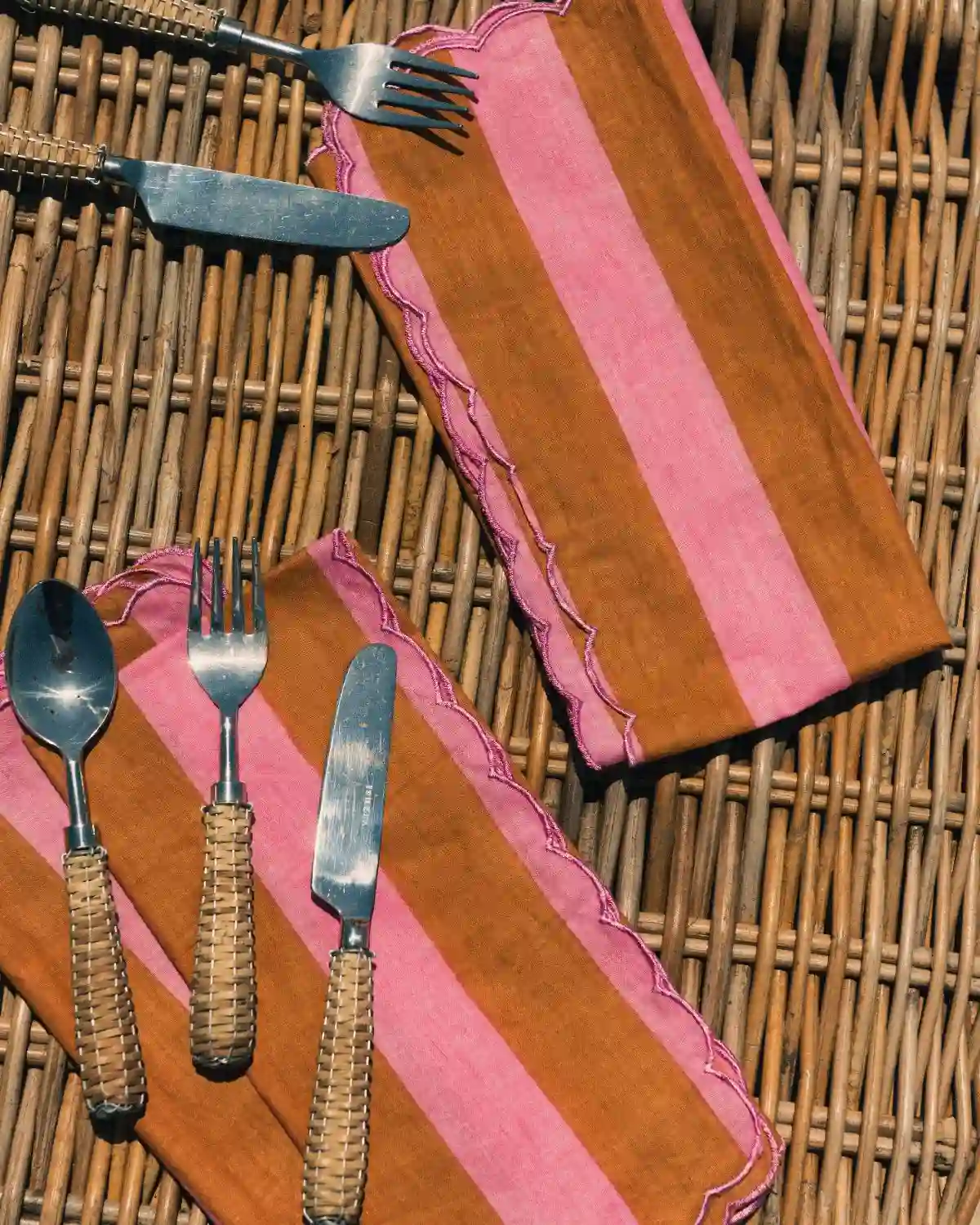 Servilletas de tela a rayas rosa y camel con borde festoneado y cubiertos de ratán.