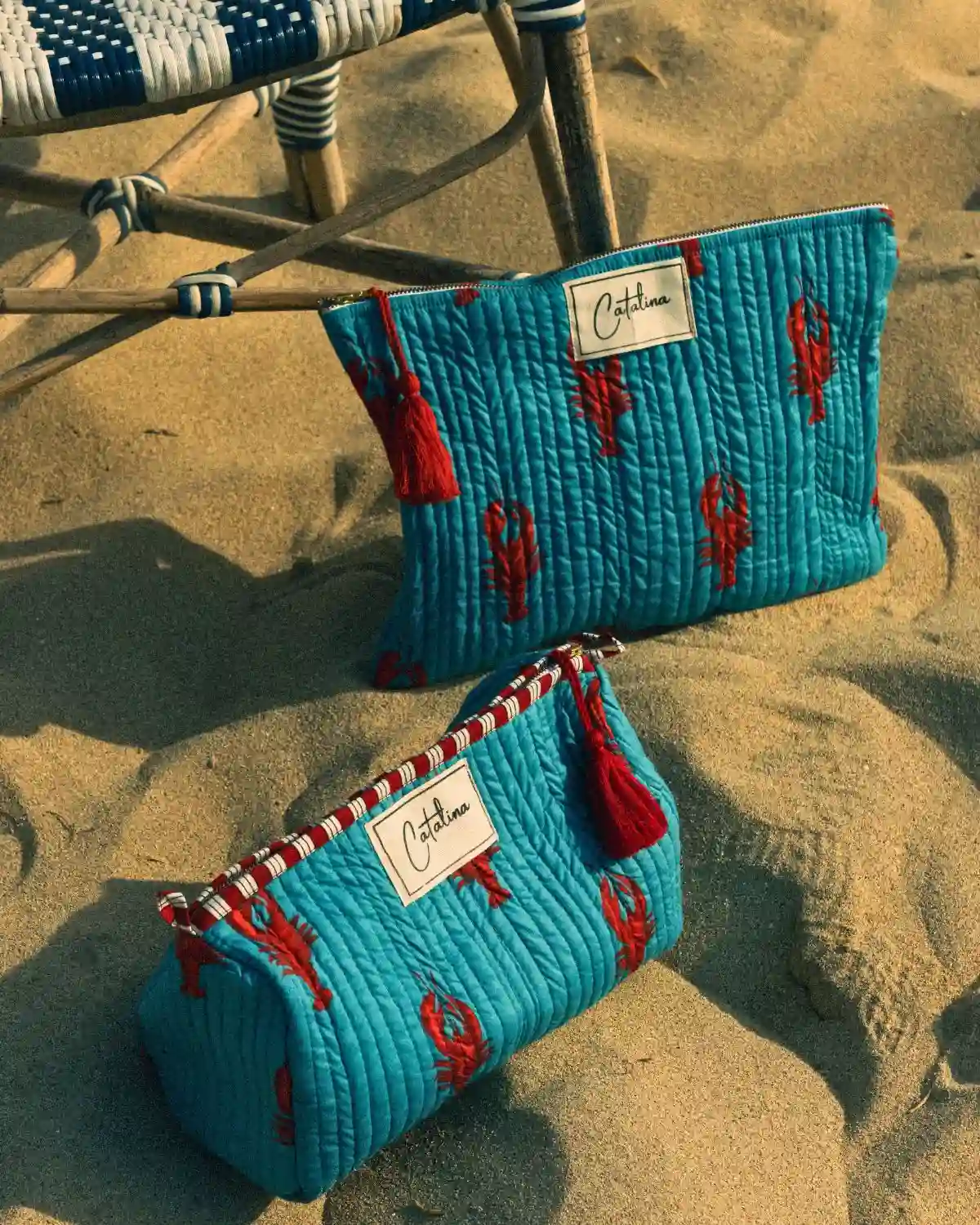 Dos formatos del Neceser Langosta Capri sobre la arena, con diseño turquesa y langostas rojas.