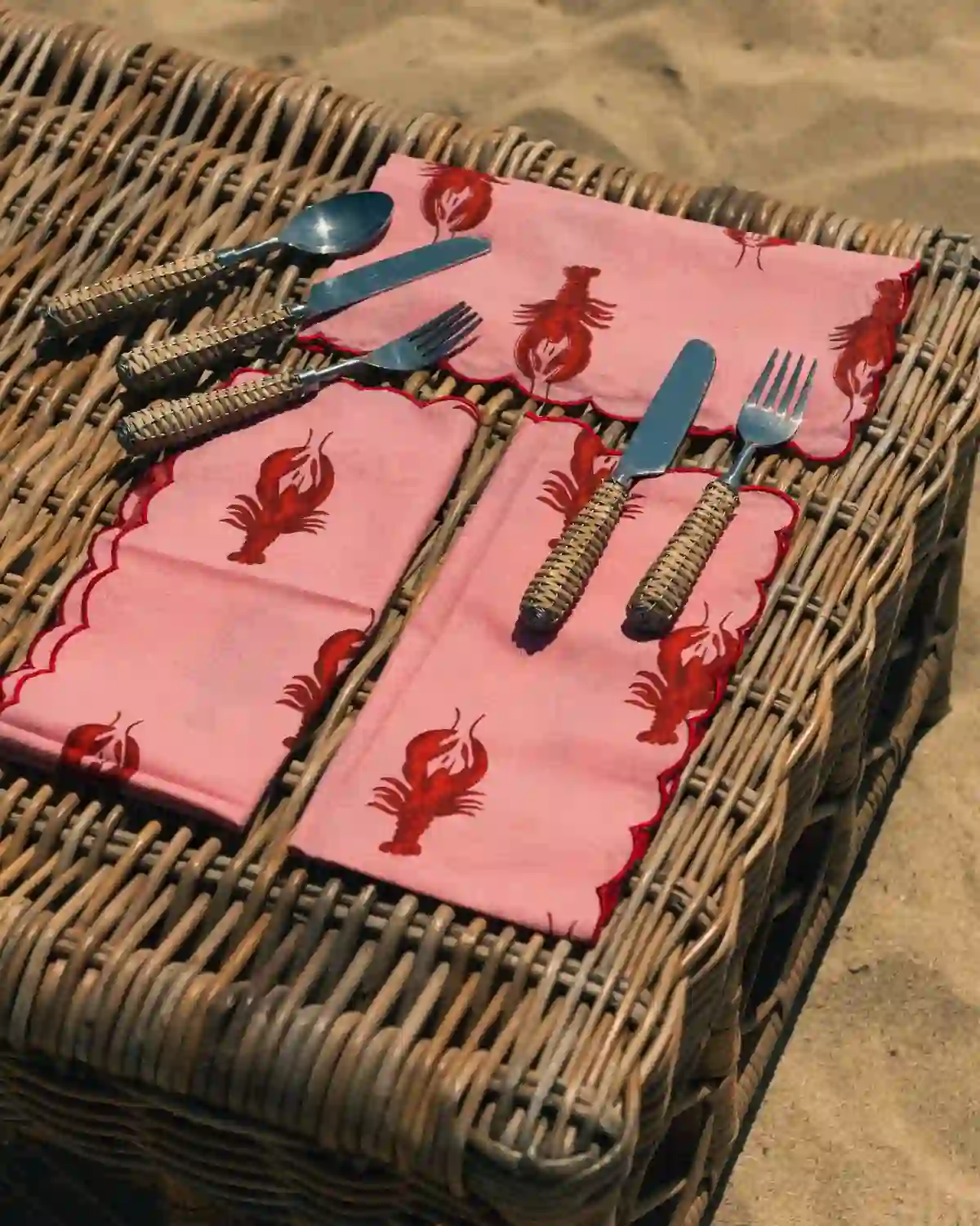 Servilletas Langostas Rosa con borde festoneado rojo, presentadas con cubiertos de ratán en una mesa de inspiración mediterránea.
