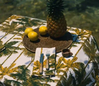 Detalle del Mantel Palmeras Mostaza de Cassai con estampado de palmeras tropicales, piña, limones y fibras naturales sobre una mesa mediterránea.