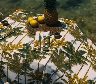 Mantel de algodón con estampado de palmeras mostaza de Cassai sobre una mesa decorada junto al mar Mediterráneo.