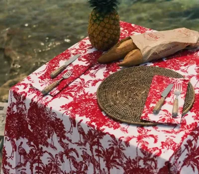 Mantel y servilletas Garza Roja de algodón de Cassai con estampado tropical en rojo y blanco sobre una mesa decorada frente al mar Mediterráneo.