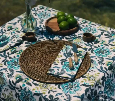 Mantel y servilletas Flor Cupido Azul de algodón de Cassai sobre una mesa decorada con flores y limas frente al mar Mediterráneo.